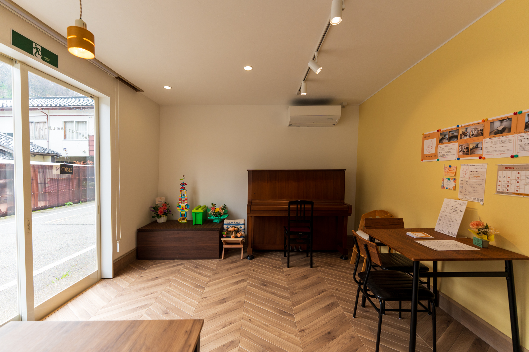 ラウンジ内観。木製テーブルと椅子、奥にピアノ。大きな窓と赤いX型の装飾窓から外の通りが見える