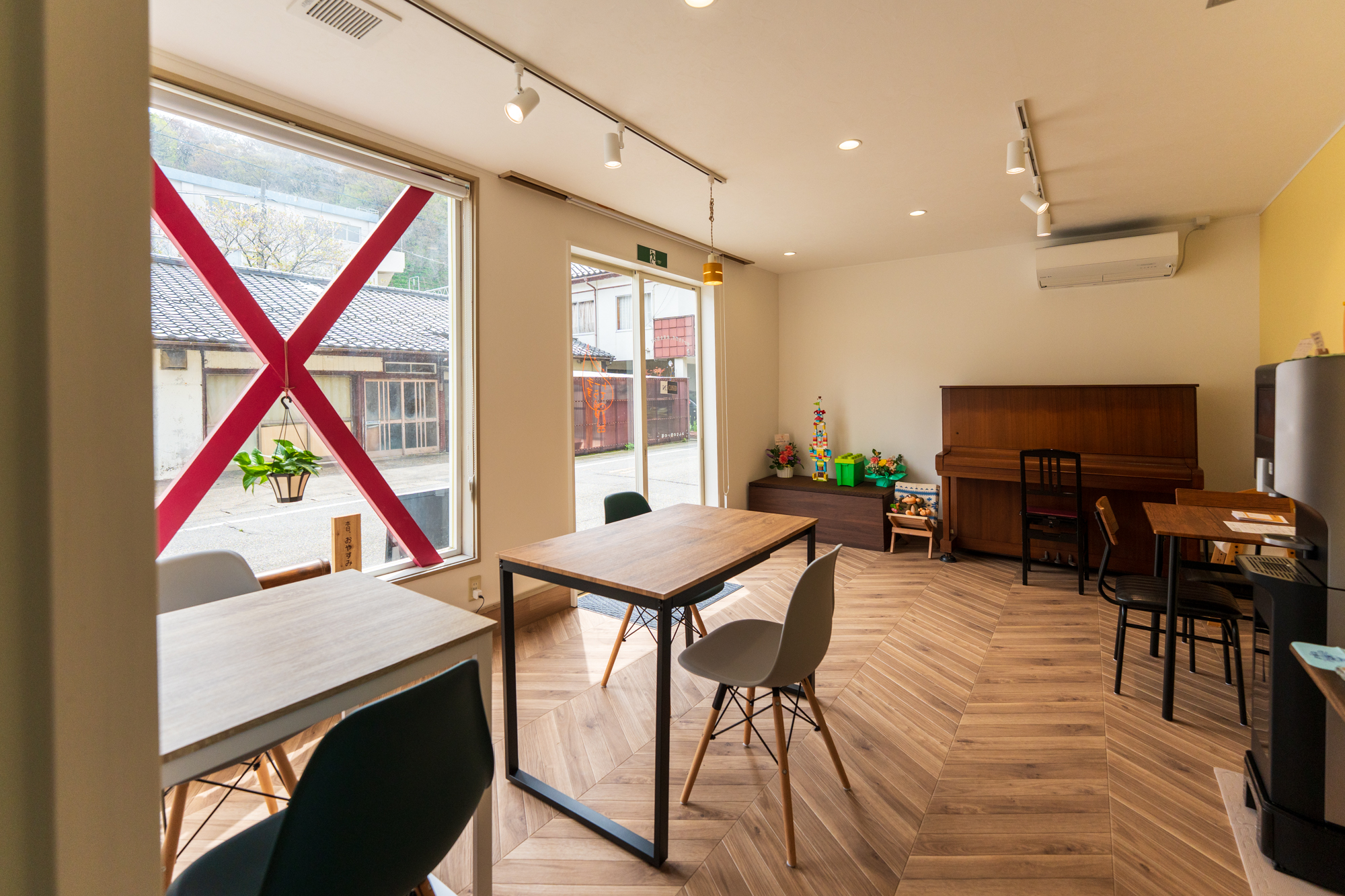 ヘリンボーン床のラウンジ全景。黄色いアクセント壁、ガラス戸から街並み、ピアノやテーブル席、コーヒーマシンなどが見える