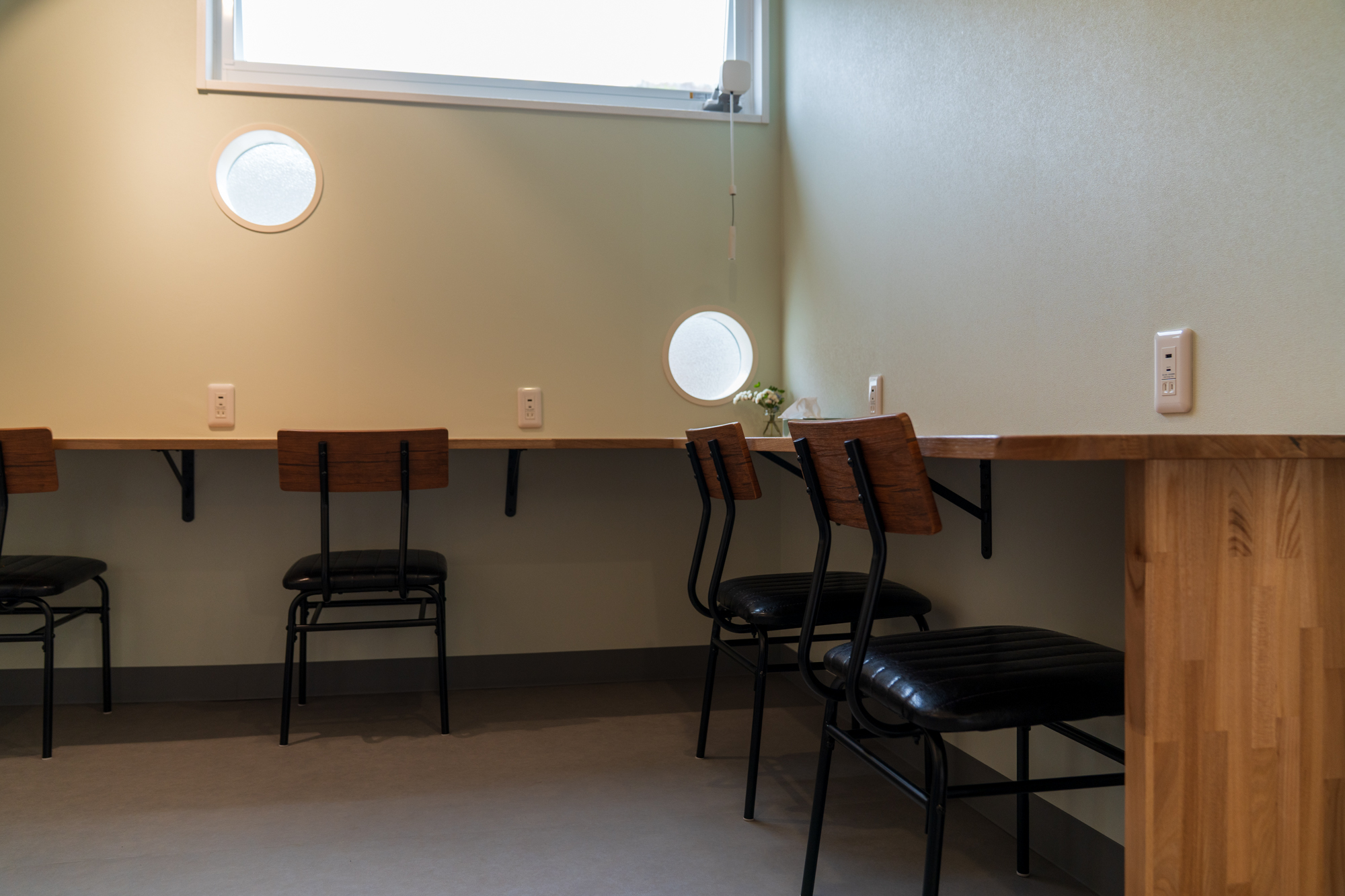 淡いグリーンの壁に沿ったL字型の木製カウンター。横長窓と丸い小窓、複数脚の椅子と壁付けコンセント